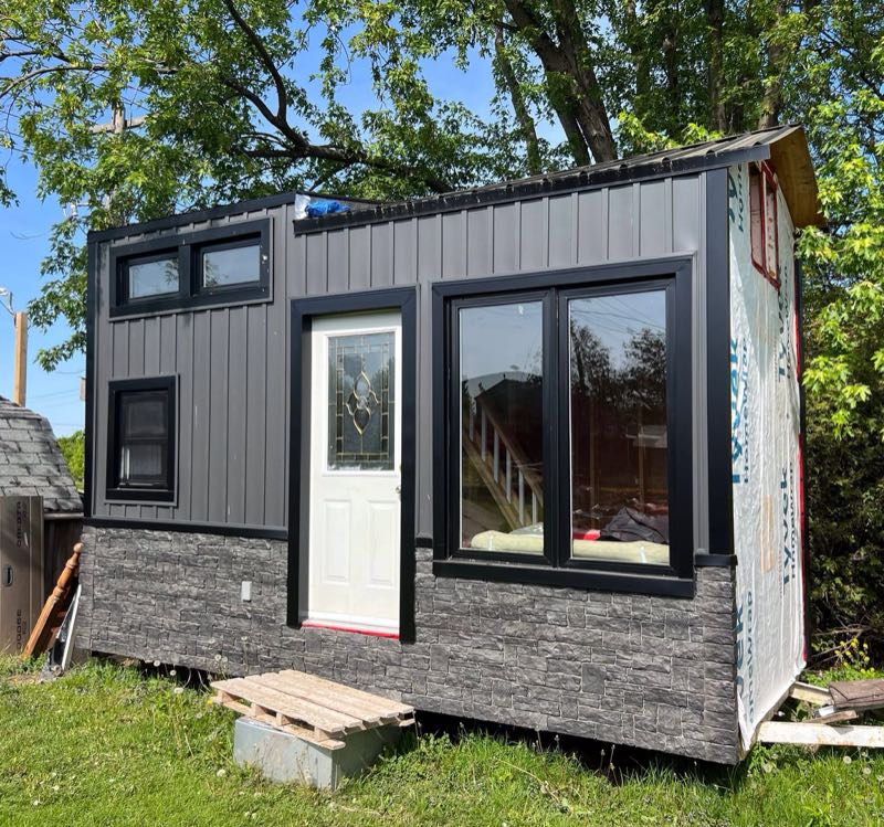 Small modern shed with dark board-and-batten siding and a stone-look base, one side still in house wrap
