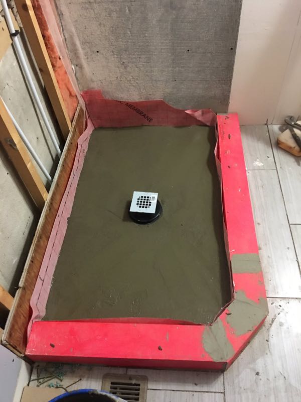 Shower base before tiling: a mortar bed sloped to a square centre drain, with red waterproofing on the curb